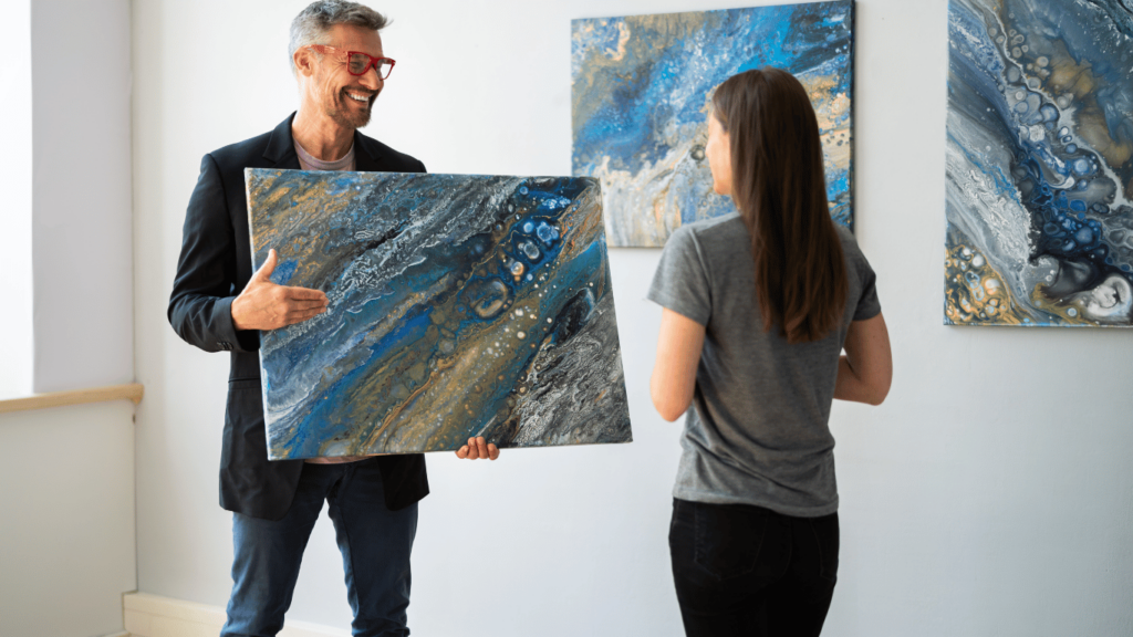 Un hombre sonriente con gafas rojas sostiene una pintura abstracta azul y dorada mientras conversa con una mujer en una galería de arte. La escena representa el interés en el arte como inversión y su potencial valor en el mercado.